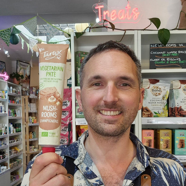A smiling man in a shop environment holds up a squeeze bottle of Tartex Mushroom Pate He is wearing a black apron with 'HEALTH & REMEDY' printed on it, and shelves of packaged goods are visible in the background.