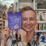 A smiling man in a black apron with a floral shirt holds up a purple bag of Crispr Bites Milk Chocolate Filled Waffle Cones on a with shelves and a pink neon sign in the background.