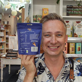 A smiling man in a tropical print shirt and black Health & Remedy apron holds up a blue bag of Crispr bites in a shop setting with shelves and a pink neon sign in the background.