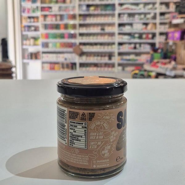 A small glass jar of spread or paste with a dark lid and tan label sits on a white counter in a blurred retail store interior.