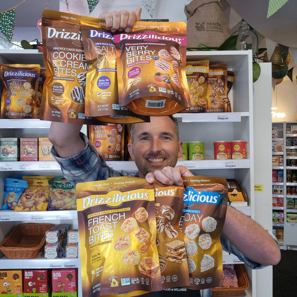 A smiling man in a store holds up multiple bags of Drizzlicious snack bites, including Cookie & Cream Bites, Very Berry Bites, French Toast Bites, and Toast Bites, with shelves of other snacks behind him.