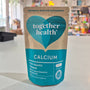 Packaging of Together Health Calcium supplements with a clear window showing the tablets inside. The package is predominantly blue and white with a heart shape design.