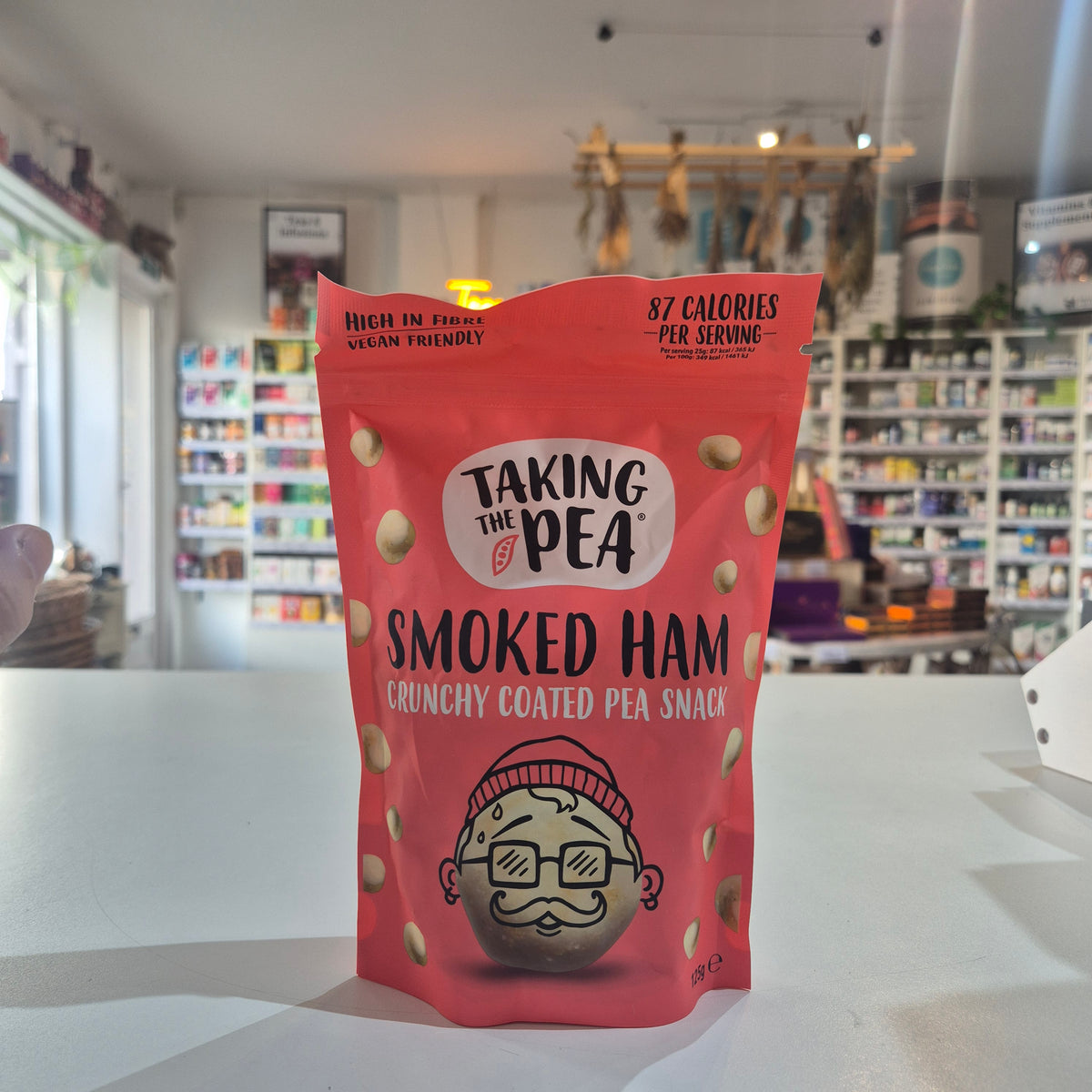 A red bag of Taking The Pea Smoked Ham Crunchy Coated Pea Snack stands on a white counter in a blurred store setting.