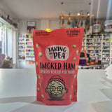 A red bag of Taking The Pea Smoked Ham Crunchy Coated Pea Snack stands on a white counter in a blurred store setting.