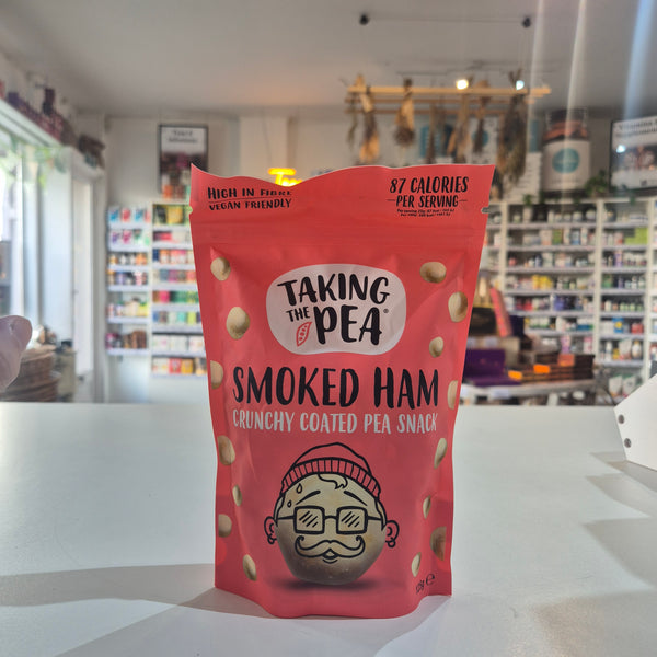 A red bag of Taking The Pea Smoked Ham Crunchy Coated Pea Snack stands on a white counter in a blurred store setting.