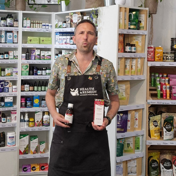 A man wearing a floral shirt and a black Health & Remedy apron stands in a shop aisle holding a small Herbetom bottle and a boxed product. Shelves behind him are stocked with various health and wellness items.