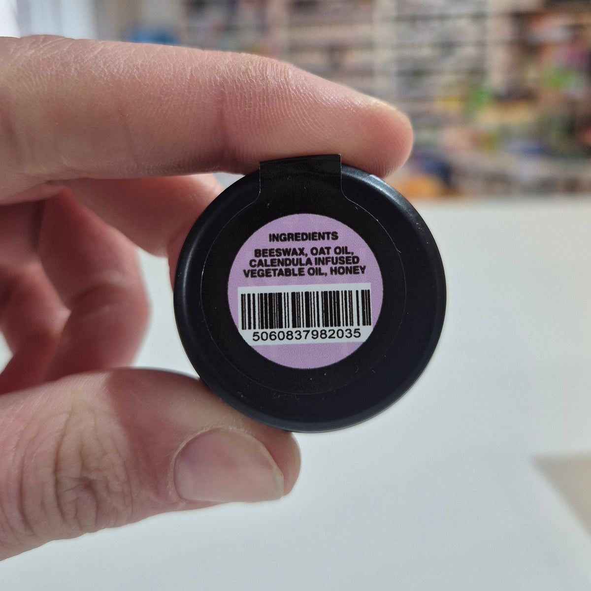 Hand holding a small container with a purple label and barcode, blurred background