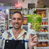 A smiling man in a shop holds up a bag of DJ&A Gourmet Snap Pea Pods. The background shows shelves stocked with various snack products and a neon 'Treats' sign.