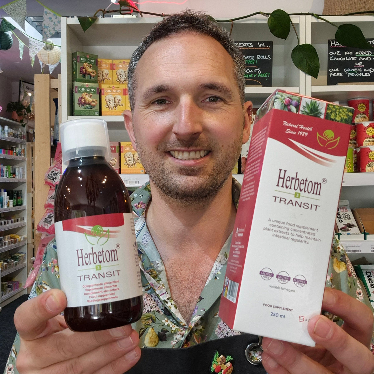 A smiling man in a floral shirt holds up a bottle and box of Herbetom Transit herbal supplement in a retail store setting.