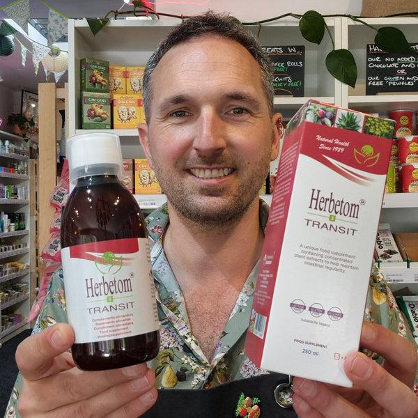 A smiling man in a floral shirt holds up a bottle and box of Herbetom Transit herbal supplement in a retail store setting.