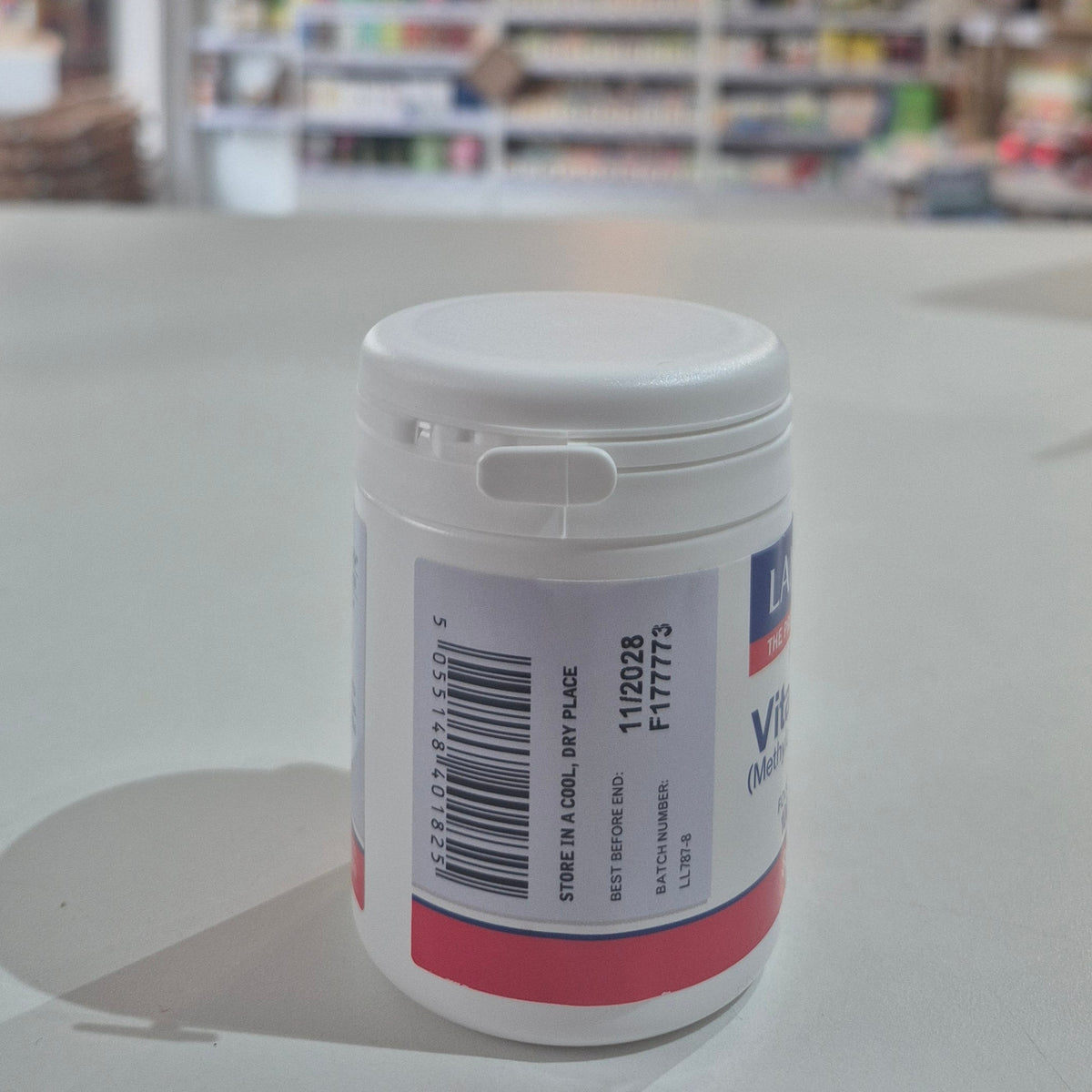 Lamberts Vitamin B12 supplement container on a table with a blurred background. White container with a label on a light gray surface, blurred background