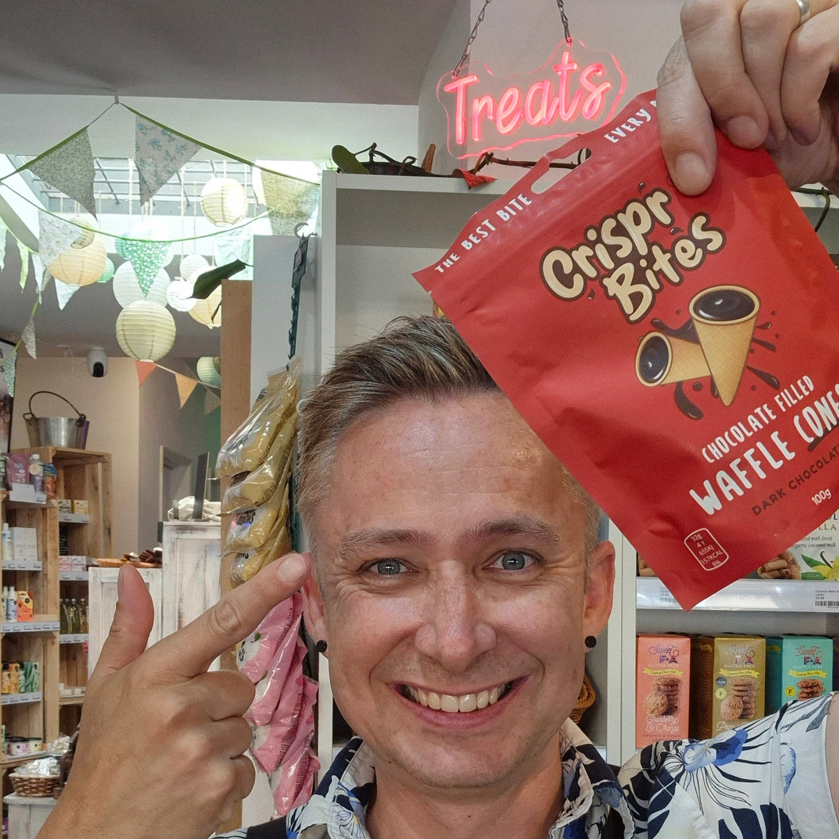 A smiling man wearing a white and navy tropical print shirt and a black 'Health & Remedy' apron holds up a red bag of 'Crisp Bites' chocolate chip waffle cookies in a shop setting.