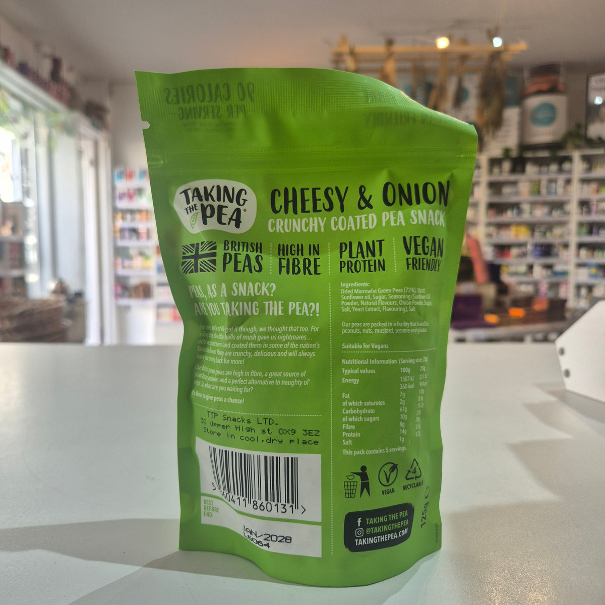 A green stand-up pouch of Taking Pea Cheesy & Onion Crunchy Coated Pea Snack, photographed on a countertop with a blurred shop interior in the background.