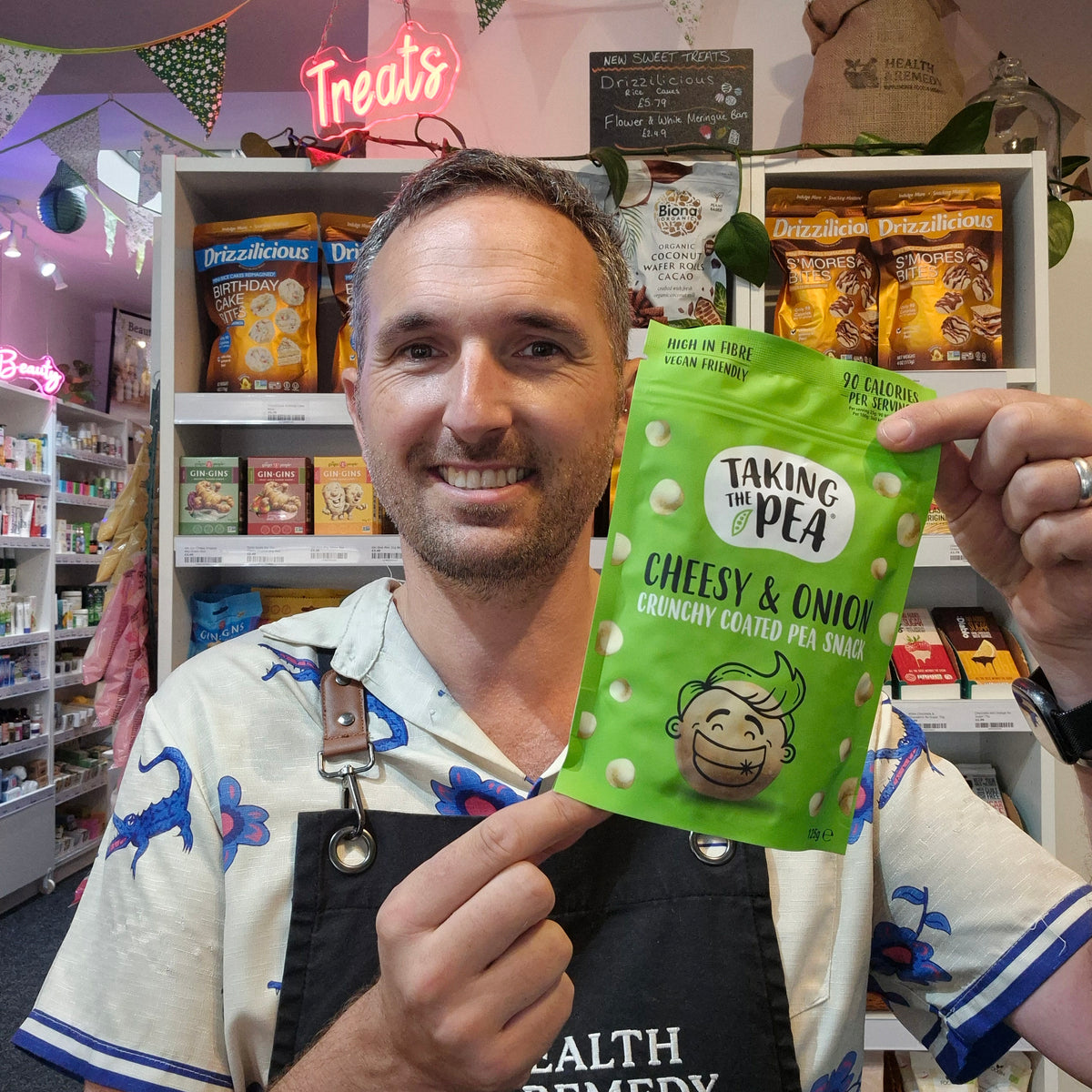 A man in a shop holding up a green bag of Taking the Pea Cheesy & 
onion Crunchy Coated Pea Snack, with shelves of other snacks in the background.