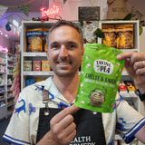 A man in a shop holding up a green bag of Taking the Pea Cheesy & 
onion Crunchy Coated Pea Snack, with shelves of other snacks in the background.