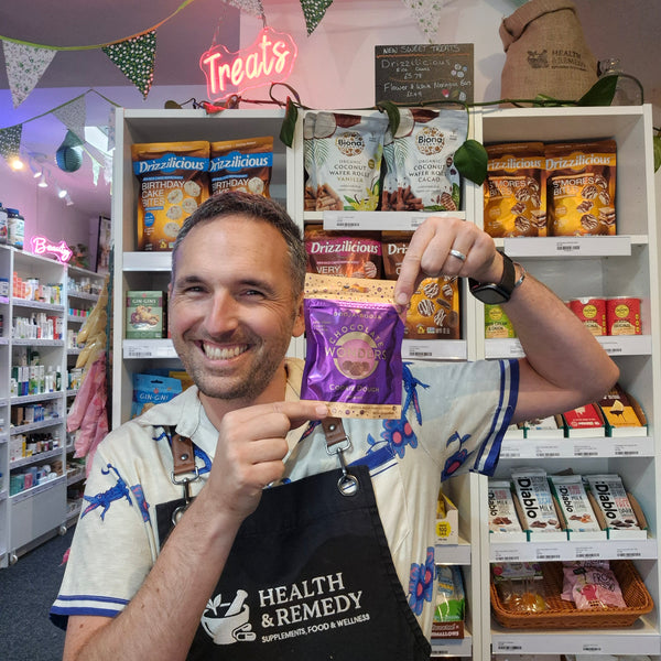 A smiling man wearing a 'Health & Remedy' apron stands in a pet store aisle, holding up a purple bag of booja booja
