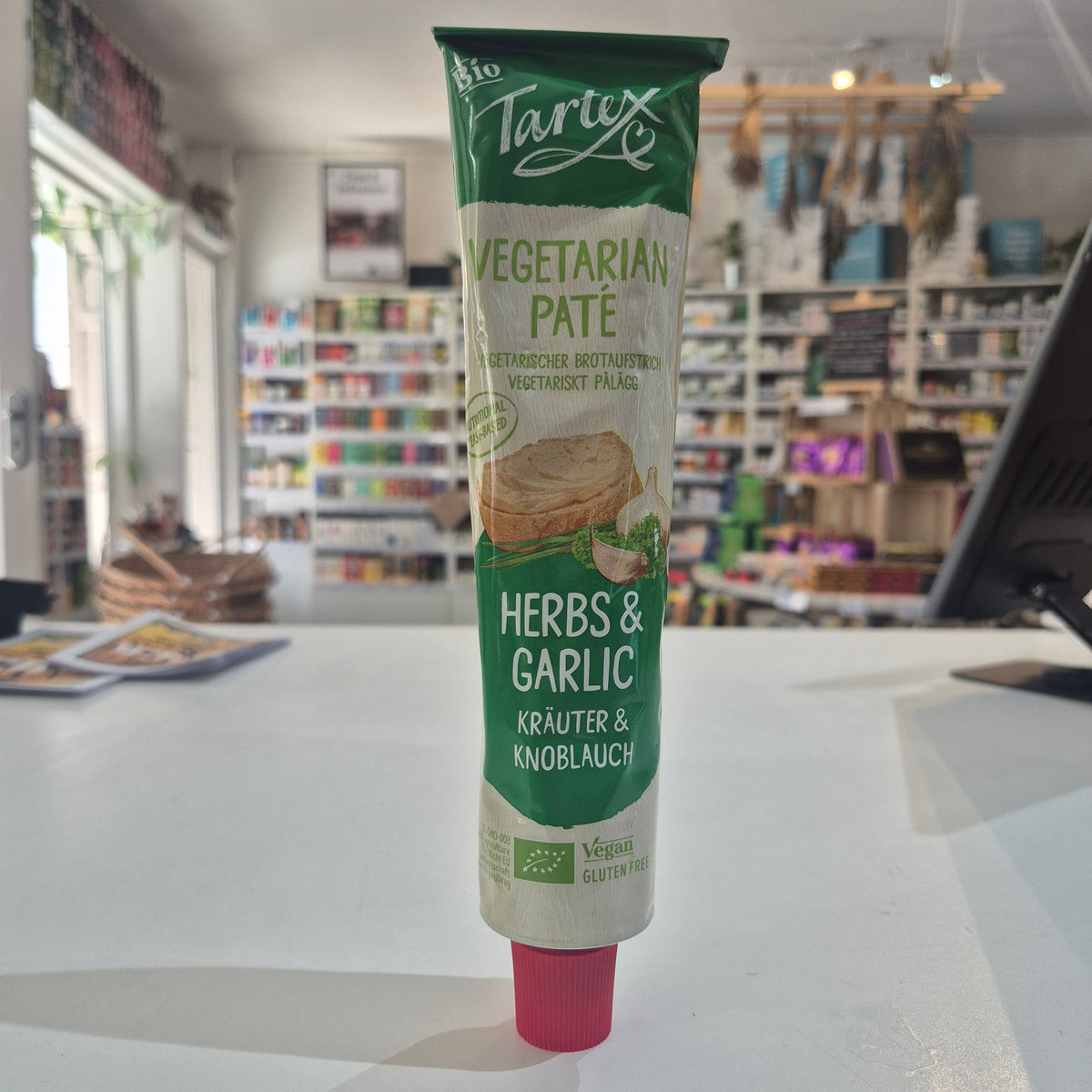 A tube of Tartex Vegetarian Pate Herbs & Garlic spread lying horizontally on a white surface, with a blurred store interior in the background.