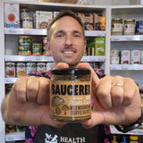 A man wearing a black Health & Remedy apron holds up a jar of Saucerer Wild Mushroom Truffle Oil in a retail store setting with shelves of other products behind him.