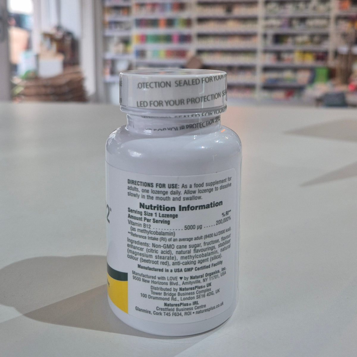 Nature's Plus Shot-O-B12 5000 Mcg lozenge in a white bottle with green and purple label, indicating wild berry flavor and dietary supplement category. Supplement bottle with nutritional information on a table in a store setting