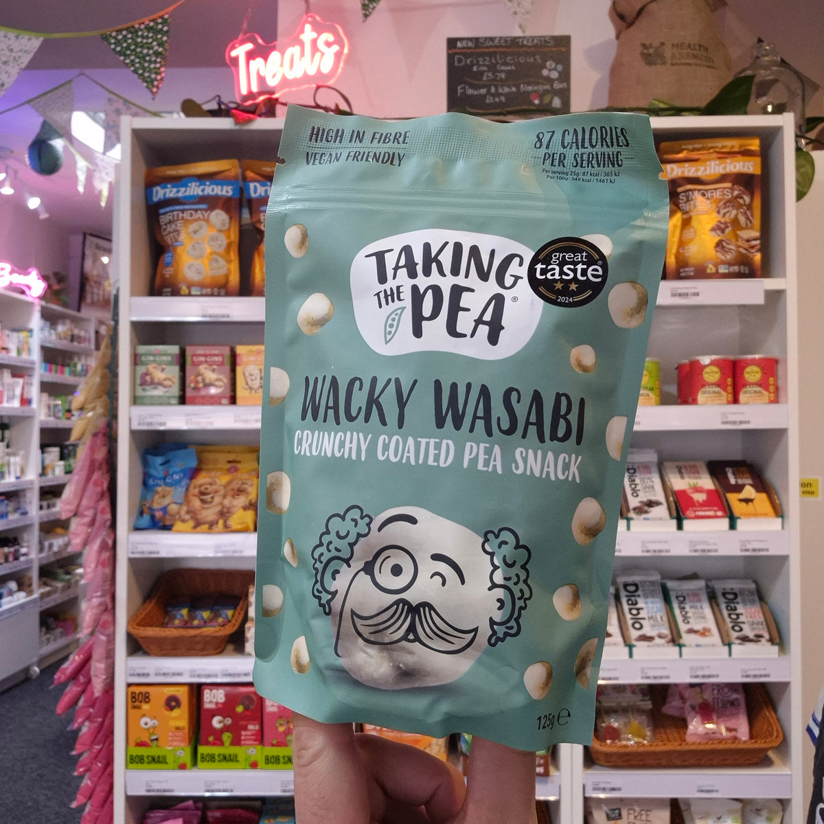 A hand holds a bag of Taking the Pea Wacky Wasabi Crunchy Coated Pea Snack in a retail store aisle. The bag is a light mint green color with white and black text and a cartoon character with a mustache.