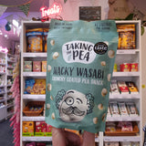 A hand holds a bag of Taking the Pea Wacky Wasabi Crunchy Coated Pea Snack in a retail store aisle. The bag is a light mint green color with white and black text and a cartoon character with a mustache.