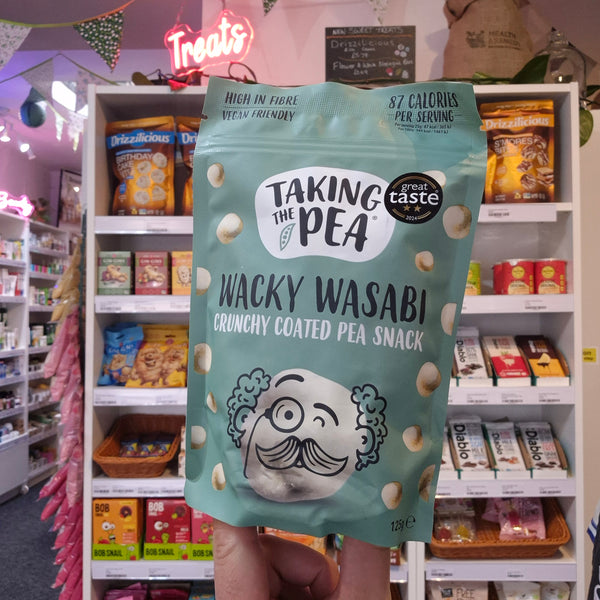 A hand holds a bag of Taking the Pea Wacky Wasabi Crunchy Coated Pea Snack in a retail store aisle. The bag is a light mint green color with white and black text and a cartoon character with a mustache.