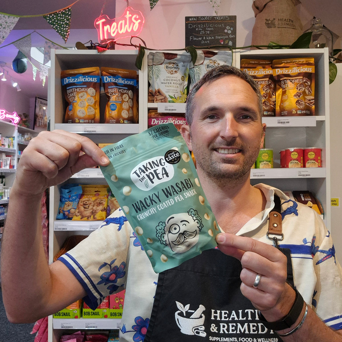 A man wearing a black Health & Remedy supplement apron holds up a mint green bag of 'Taking the Ple' Wacky Wasabi! Crunchy Coated Peanuts in a retail store setting with shelves of snacks behind him.