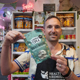 A man wearing a black Health & Remedy supplement apron holds up a mint green bag of 'Taking the Ple' Wacky Wasabi! Crunchy Coated Peanuts in a retail store setting with shelves of snacks behind him.