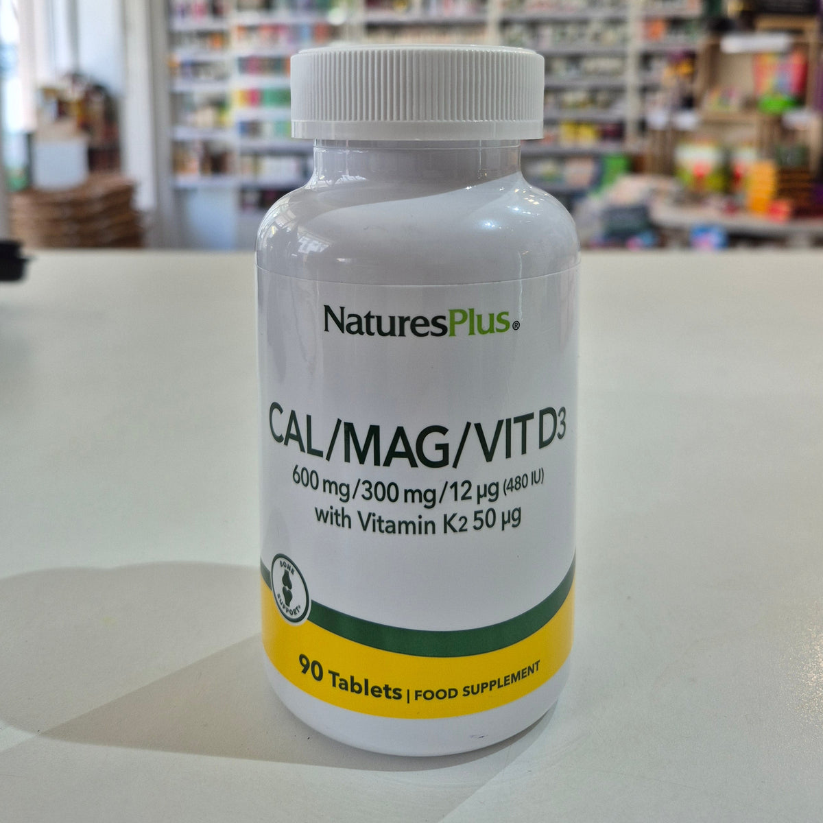 NaturesPlus Cal/Mag/Vit D3 supplement bottle on a table with a blurred store background