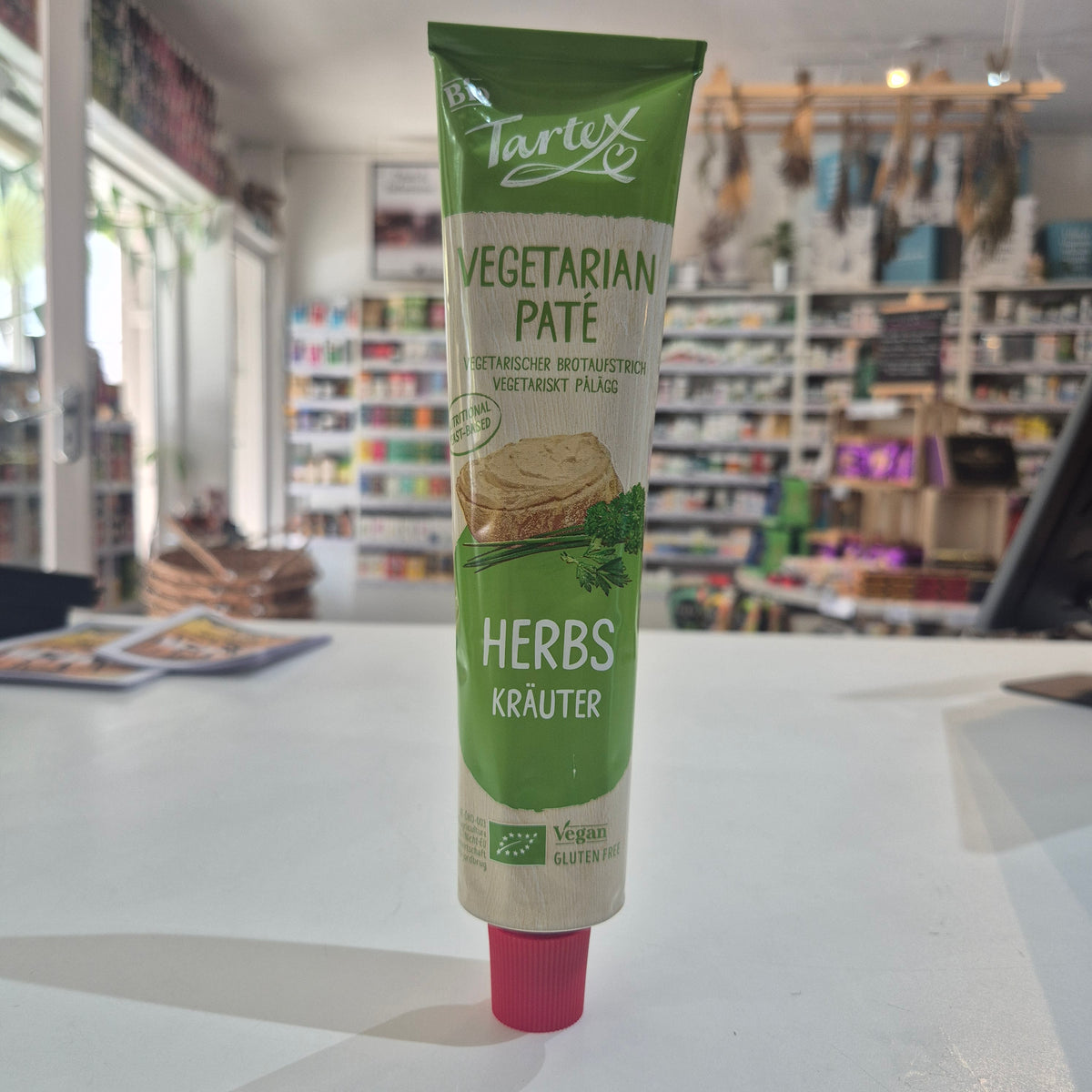 A tube of Tartex Vegetarian Pate with Herbs featuring a green and beige label with a pink cap, shown against a blurred background of a store interior.