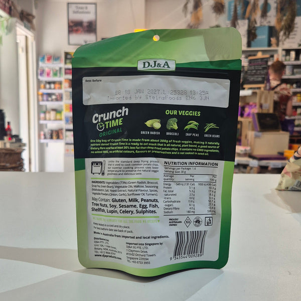 A stand-up pouch of DJ&A Crunch Time organic vegetable chips placed on a white countertop inside a retail store. The back of the green and black package is visible, with store shelves and fixtures blurred in the background.