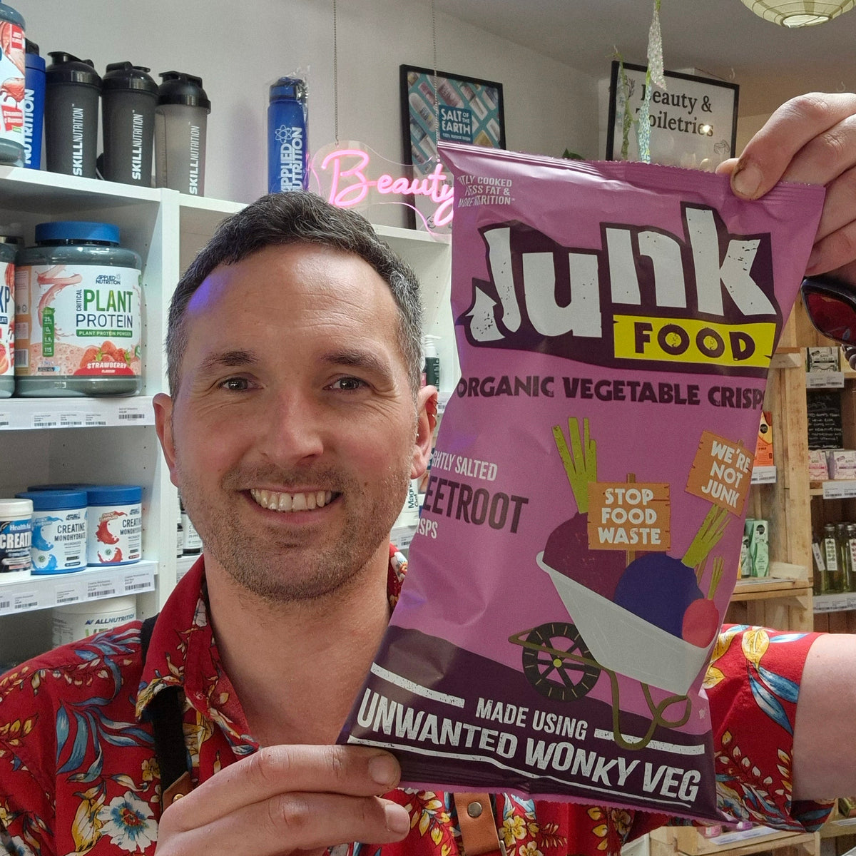 Man holding a pink 'Junk Food' organic vegetable crisp packet in a store setting.