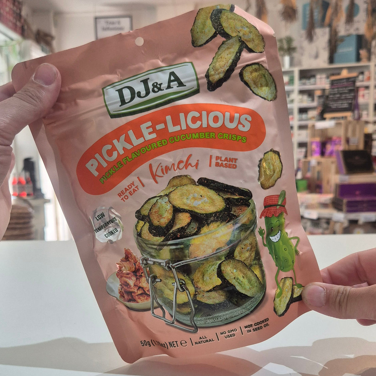 A person holds up a bag of Dj&A Pickle-Licious Kimchi Pickled Zucchini slices in a brightly lit store setting.