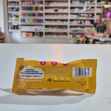A packaged meringue bar snack placed on a white counter in a blurred shop interior with shelves and hanging decor in the background. The back of a yellow and white patterned hair extension packaging sleeve, showing text instructions and a barcode, resting on a white surface with a blurred beauty store background.