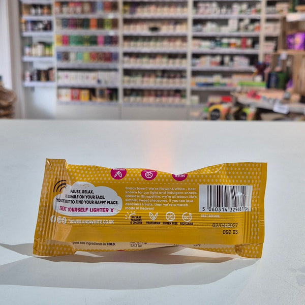 A packaged meringue bar snack placed on a white counter in a blurred shop interior with shelves and hanging decor in the background. The back of a yellow and white patterned hair extension packaging sleeve, showing text instructions and a barcode, resting on a white surface with a blurred beauty store background.