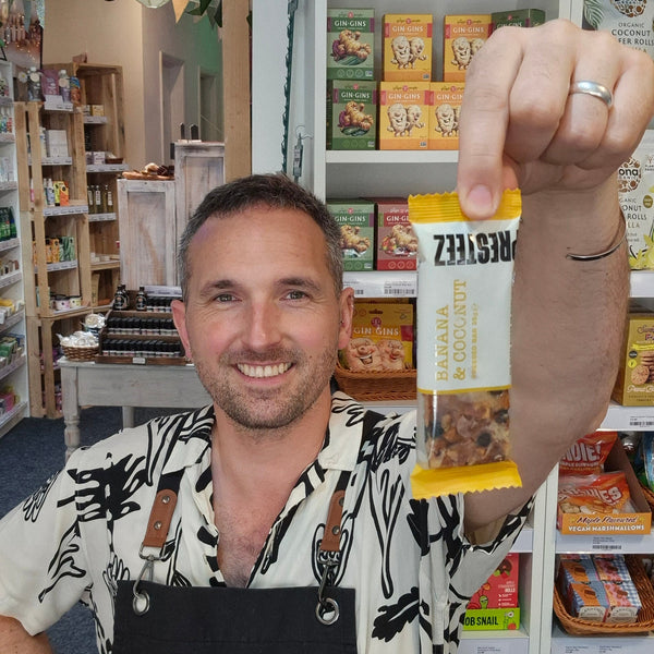A smiling man in a black Health & Remedy apron holds up a packaged snack bar inside a retail shop. The package is yellow and white with the brand name Presteez visible.