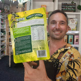 A smiling man in a yellow tropical shirt holds up the back of a green and yellow DJ&A Jalapeno Pickle-Licious snack pouch in a retail shop setting.